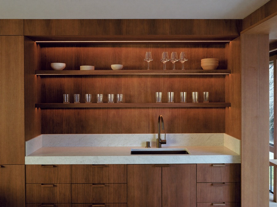 Home bar in dark wood with a thick white veined counter, a dark faucet, and lit open shelves holding glasses and bowls.