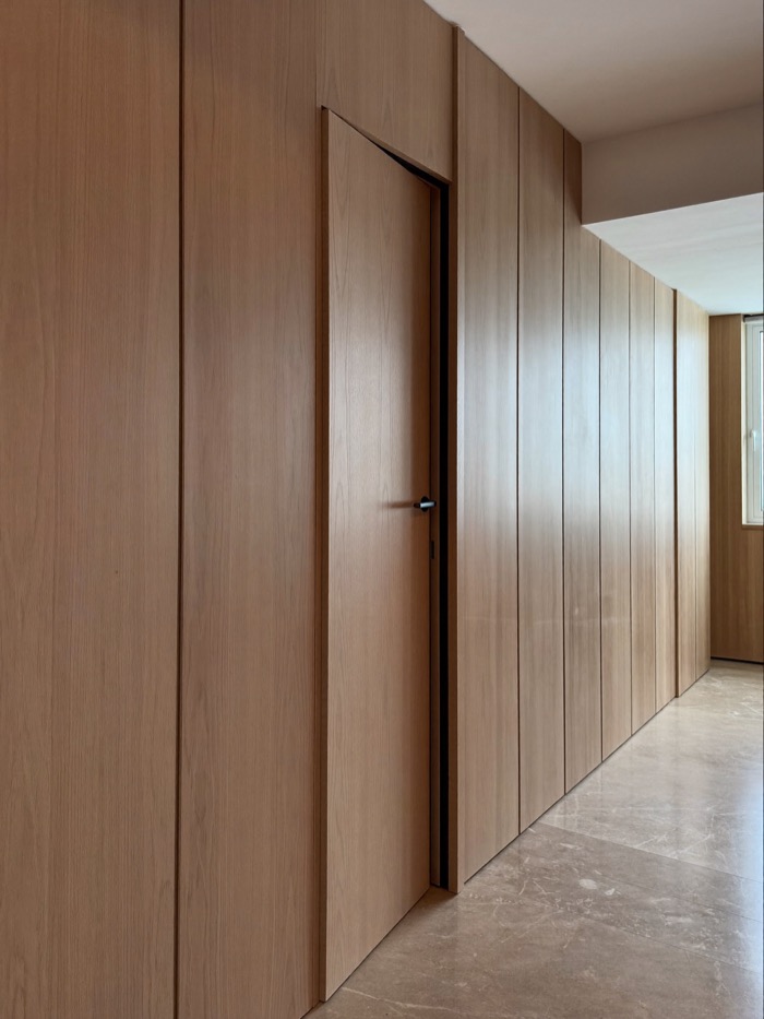 Corridor lined with full-height pale wood panels with narrow reveals, one panel a flush door standing slightly open.