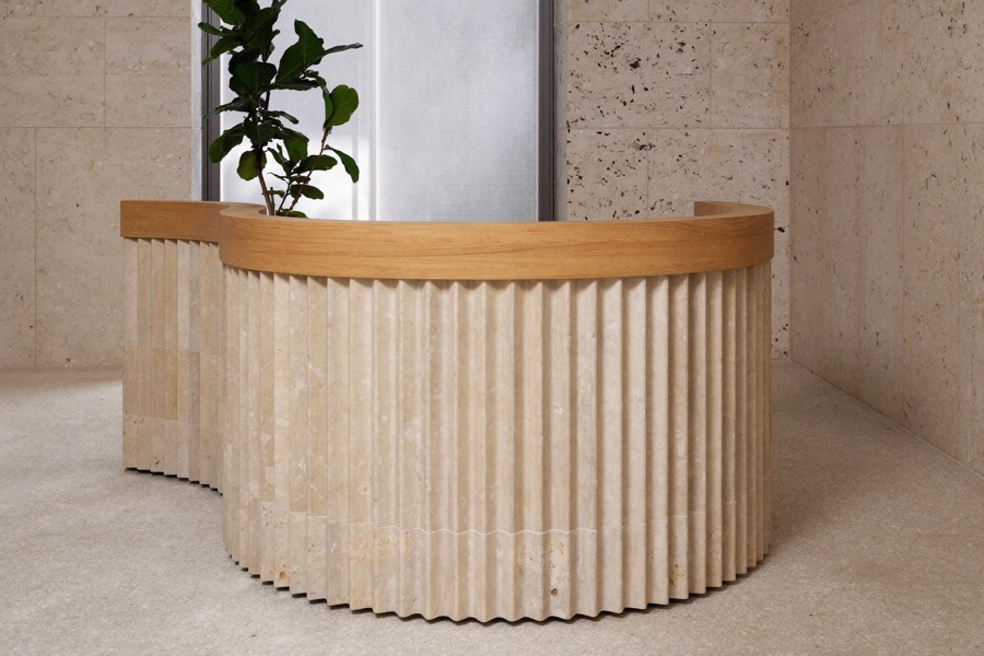 Curved reception desk with a fluted pale front and a warm wood top cap, in a pale stone lobby.