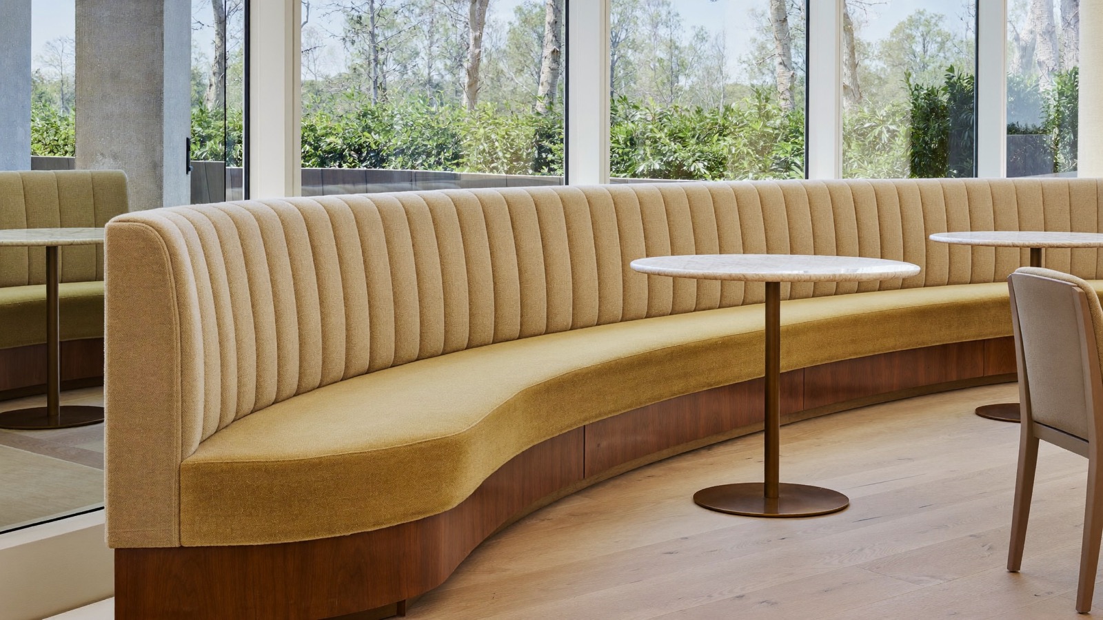 Long curved channel-stitched banquette on a wood base beside tall windows, with small round pale-topped tables.