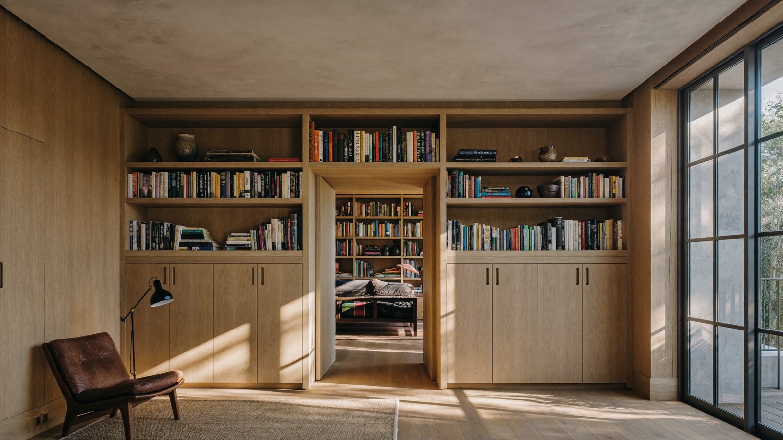Home library with floor-to-ceiling pale wood shelves and cabinets around a doorway, in late afternoon light.