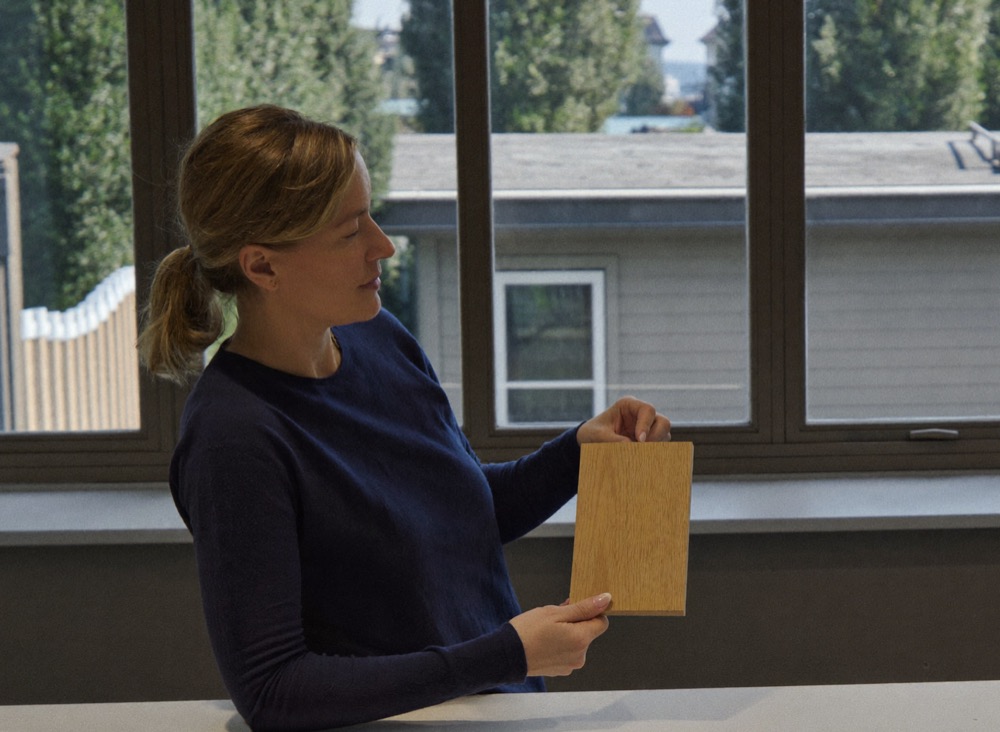 A woman in a navy sweater holds a pale wood finish sample up to the window light, studying its tone.