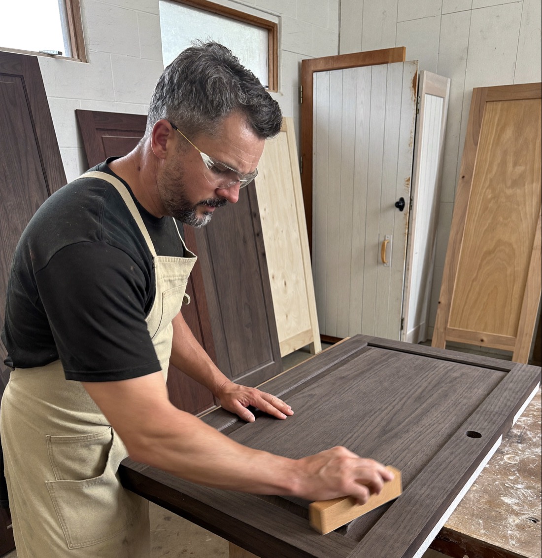 A woodworker in safety glasses and a canvas apron sands a dark wood cabinet door with a block at the workbench, with finished doors leaning against the wall behind him.
