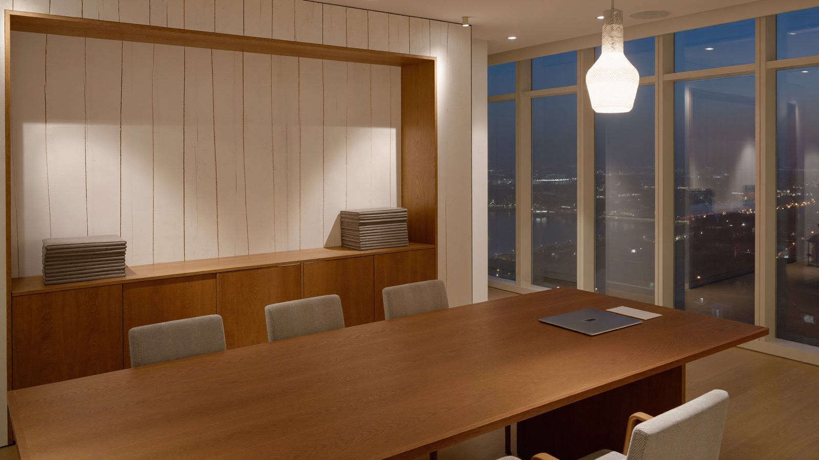 Boardroom at dusk with a long wood table and a built-in wood credenza, furniture pads stacked on the credenza, city lights through tall windows.